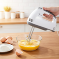 White grey electric hand mixer whisking eggs in glass bowl on wooden kitchen counter lifestyle